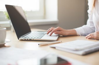 Pessoa escrevendo num macbook com um bloco de notas do lado - Europass