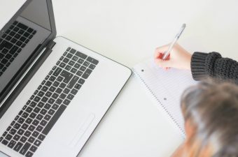 Uma pessoa escrevendo em um caderno com um lápis, e um notebook do lado