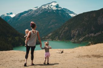 Uma mulher com uma criança no colo e outra lhe dando a mão, olhando para uma paisagem que tem um lago e, ao fundo, uma montanha