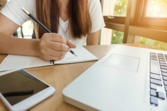 Menina escrevendo em um caderno, com um notebook à sua frente e um celular ao lado