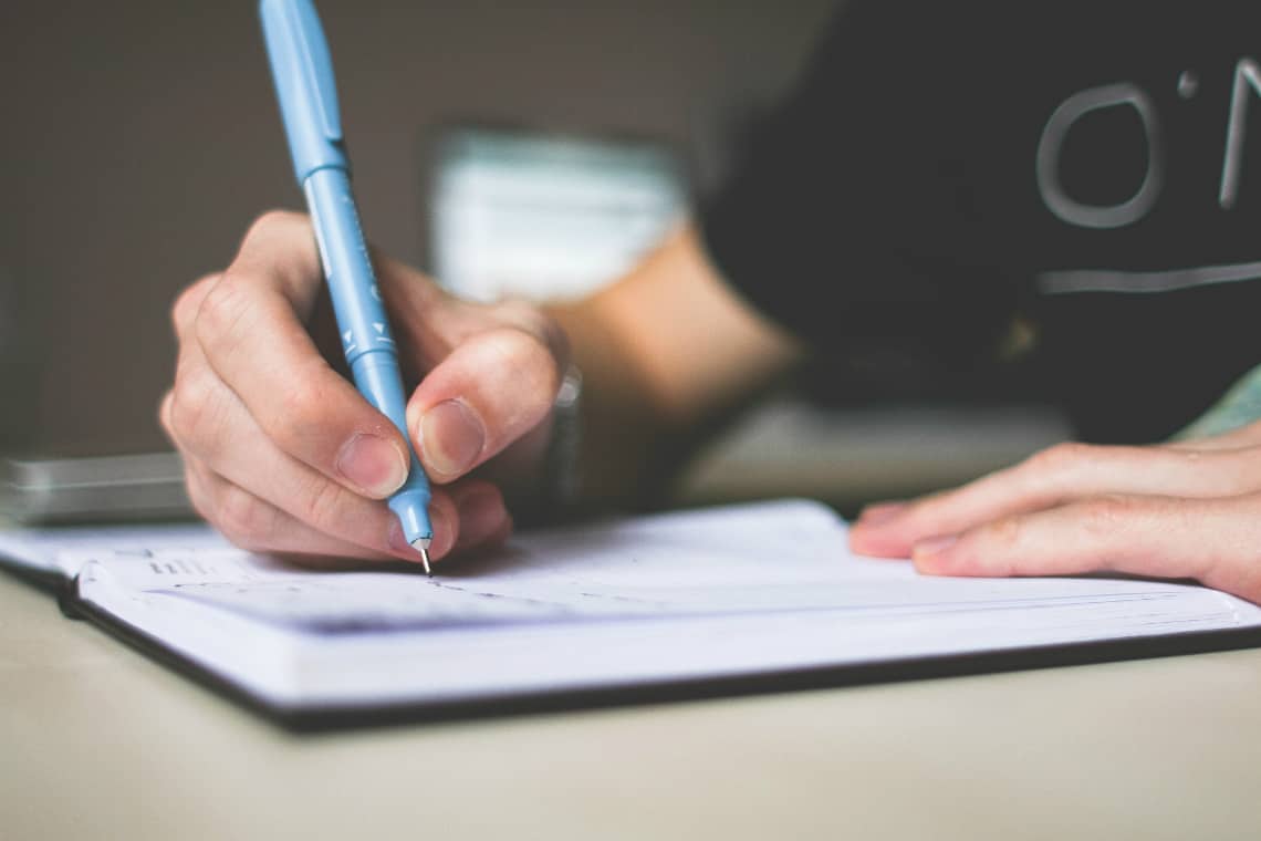 Pessoa escrevendo em um caderno com uma caneta azul, redação, writing, essay, estudar nas férias