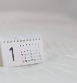 Um calendário pequeno branco com as letras pretas e indicando o número 1 como dia da semana em cima de uma mesa branca que completa todo o fundo da imagem.