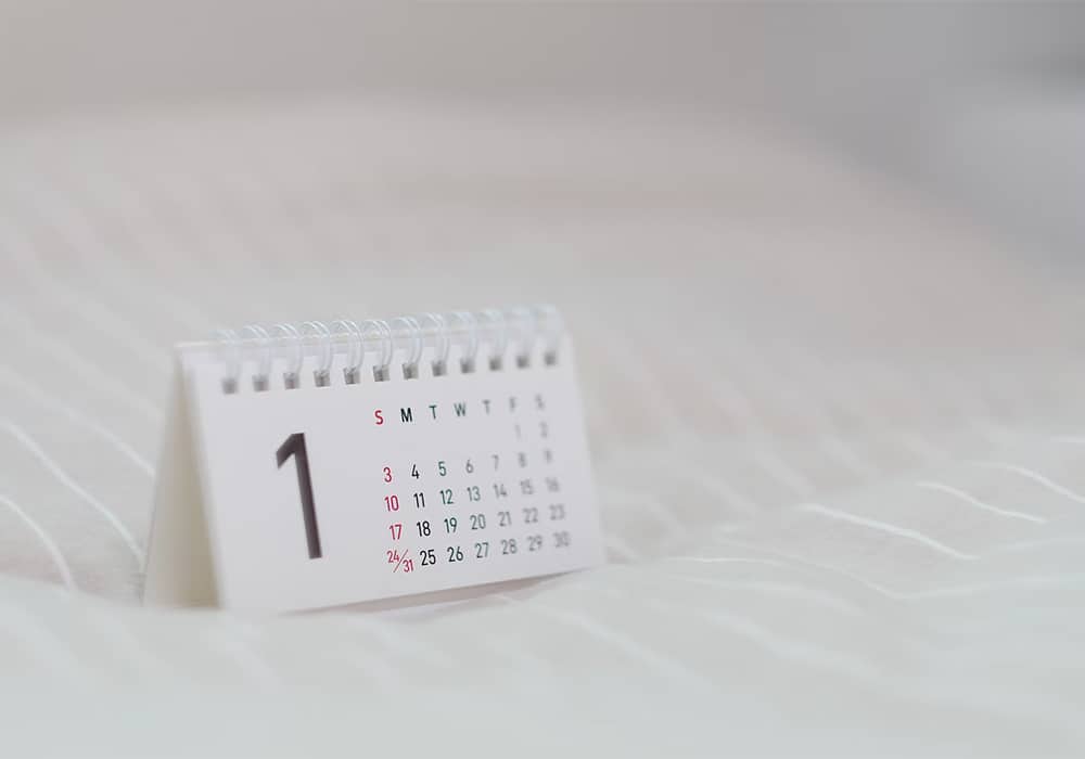 Um calendário pequeno branco com as letras pretas e indicando o número 1 como dia da semana em cima de uma mesa branca que completa todo o fundo da imagem.