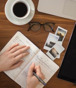 Pessoa escrevendo num caderno sobre uma mesa com fotos, cadernos, um computador e café - essay