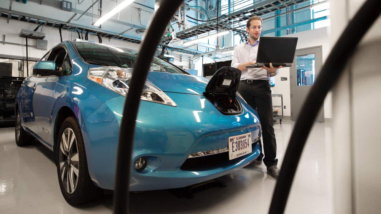 Pessoa com notebook em pé ao lado de um carro elétrico que está sendo carregado - Mestrado em carros elétricos