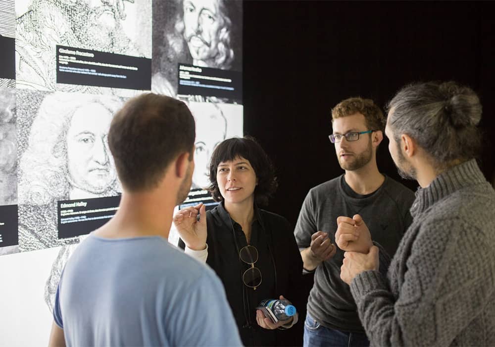 4 pessoas (3 homens e uma mulher) conversando em pé ao lado de diversas telas em preto e branco com rosto de pintores e obras artísticas