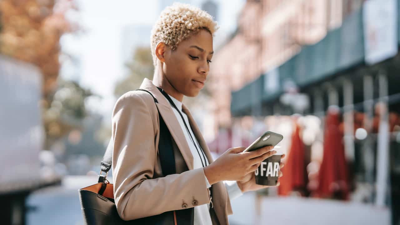 Moça segurando o celular com uma mão no meio da rua, com um café na outra - falantes nativos