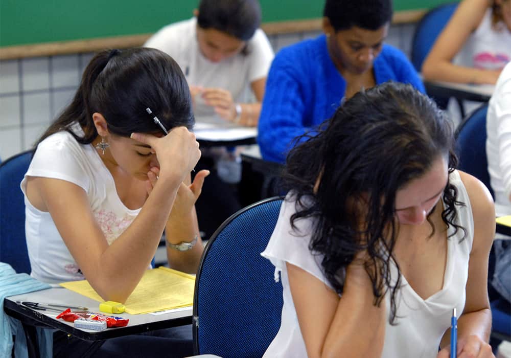 Duas estudantes sentadas em cadeiras escolares dentro de uma sala de aula e concentradas realizando provas.