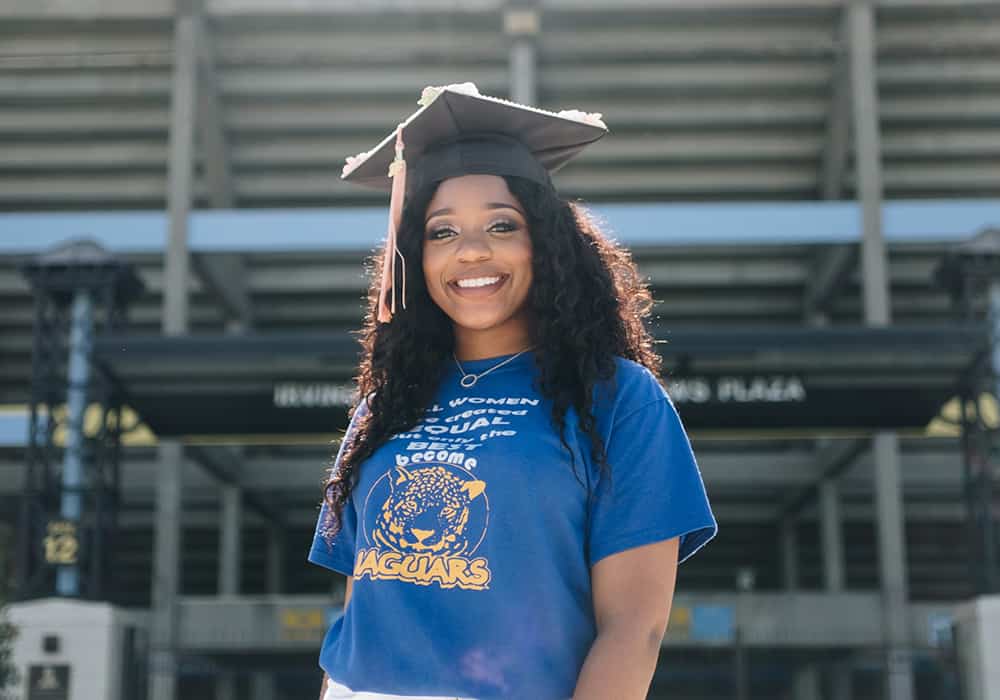 Moça no centro da imagem com chapéu de formatura e uma camiseta com estampa universitária. Ao fundo tem a parte de trás de uma arquibancada.