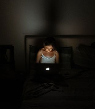 Menina usando um notebook sentada na cama em um quarto escuro