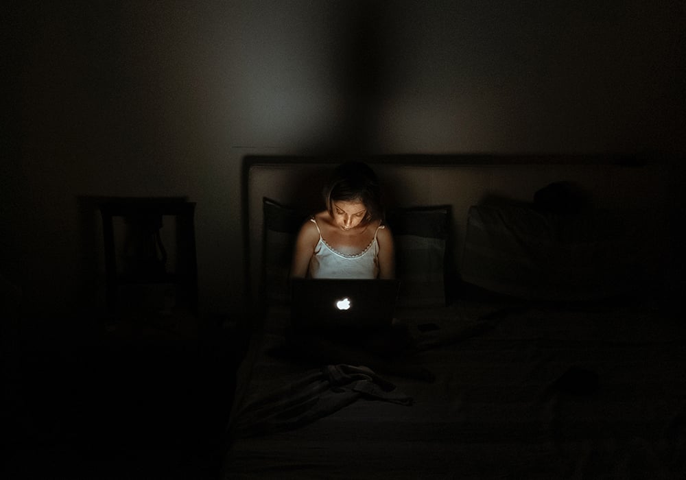 Menina usando um notebook sentada na cama em um quarto escuro