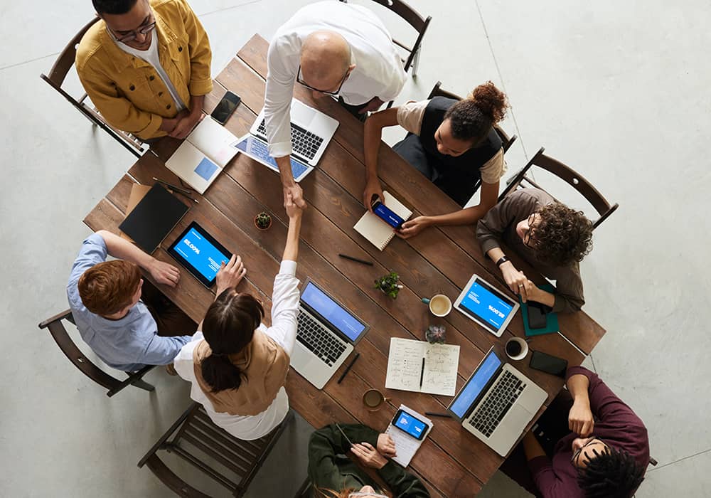 Vista de cima de 8 pessoas conversando e sentadas ao redor de uma mesa de madeira com computadores e tablets ligados em cima.