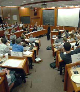 Sala de aula da harvard Business School tipo plateia circular com todas as cadeiras cheias e uma professora falando ao centro. No fundo, há três lousas, duas verdes nas beiradas e uma branca no centro.
