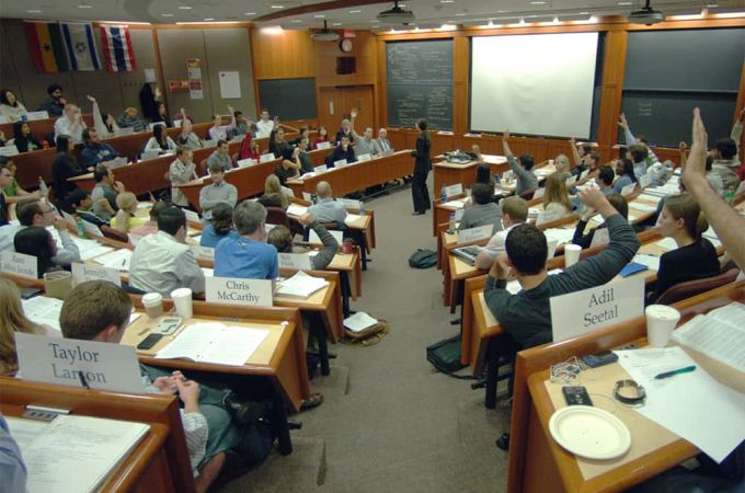 Sala de aula da harvard Business School tipo plateia circular com todas as cadeiras cheias e uma professora falando ao centro. No fundo, há três lousas, duas verdes nas beiradas e uma branca no centro.