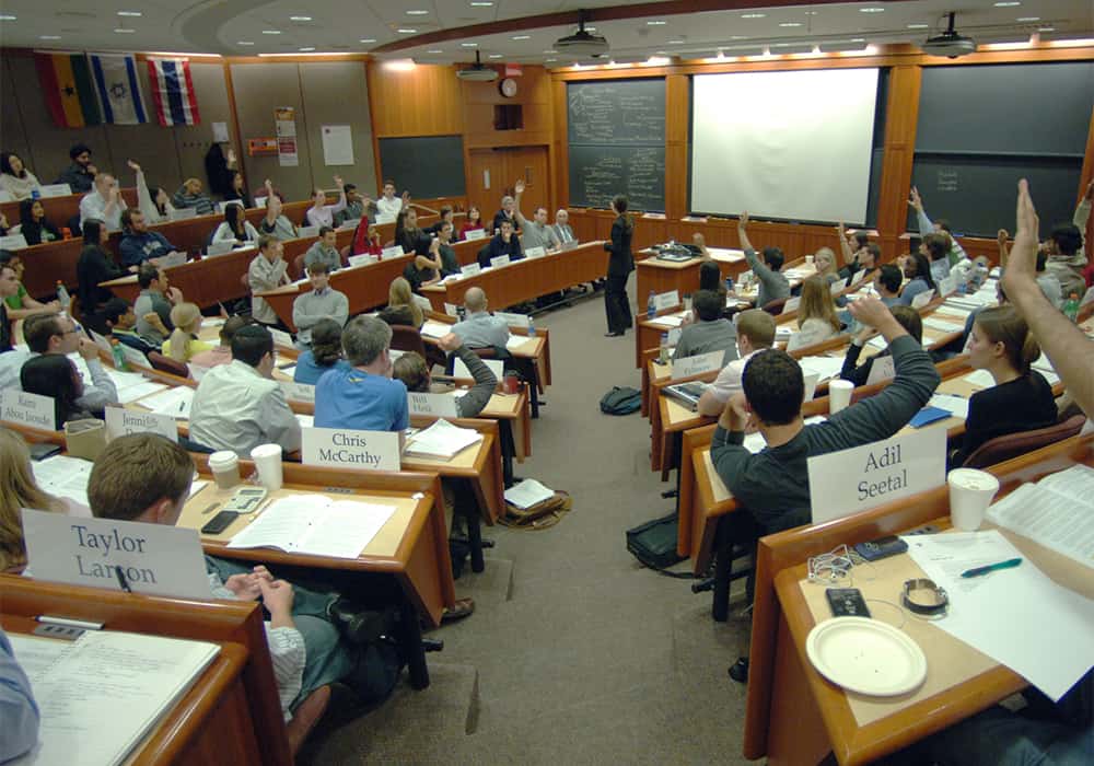 Sala de aula da harvard Business School tipo plateia circular com todas as cadeiras cheias e uma professora falando ao centro. No fundo, há três lousas, duas verdes nas beiradas e uma branca no centro.