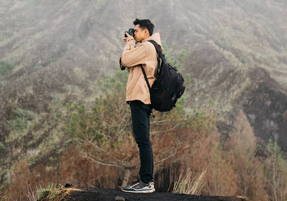 homem em pé fotografando algo a frente, no fundo uma cadeia de montanhas com vegetação baixa