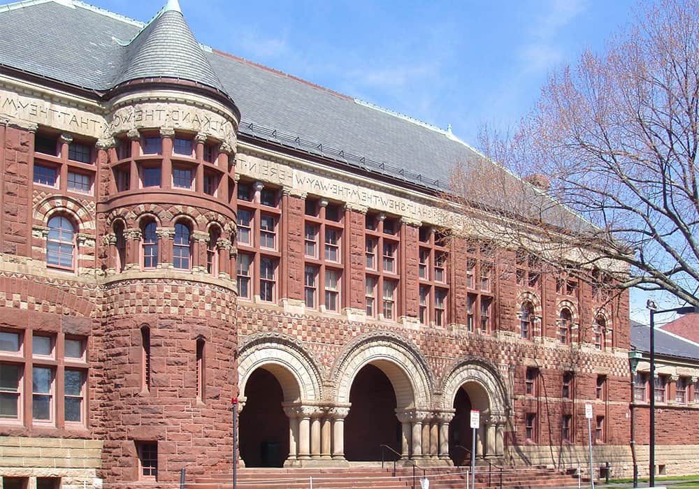 Entrada do Austin Hall da Faculdade de Direito de Harvard. O prédio é construído com tijolos antigos alaranjados e uma entrada com três grandes arcos.