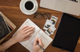 Pessoa escrevendo num caderno sobre uma mesa com fotos, cadernos, um computador e café - essay