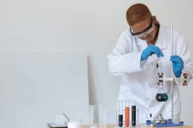Moça de jaleco, óculos e luvas de plástico mexendo em equipamentos de laboratório - science ambassador